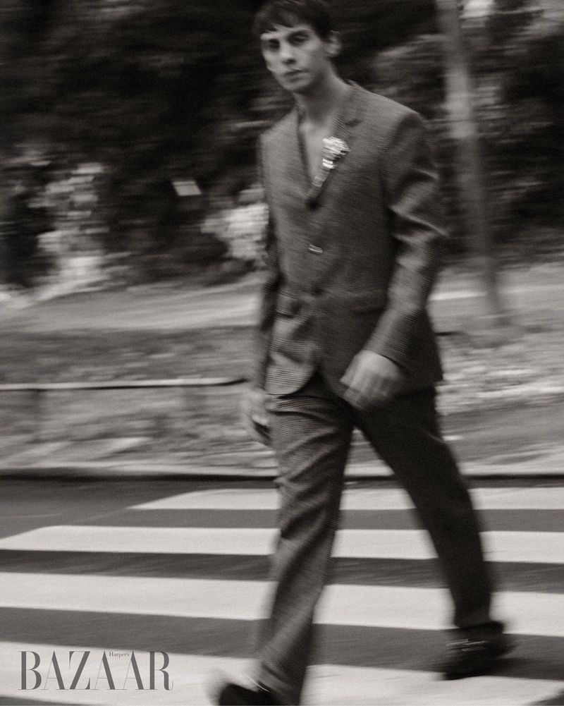 Monochrome shot of a slender man in a suit walking across a zebra crosswalk with blurred trees, capturing a fashion editorial style.