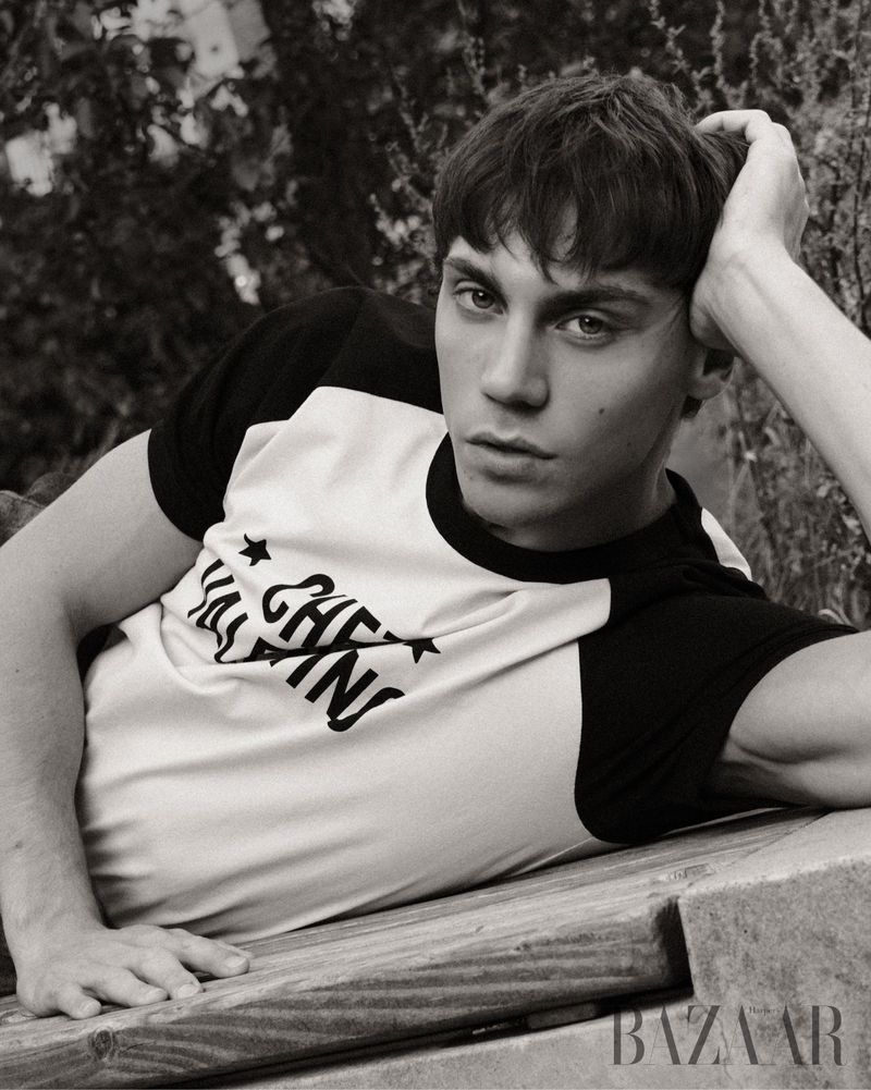 Black and white portrait of a young adult man with dark hair, wearing a short-sleeved t-shirt, lying outside on a wooden surface.