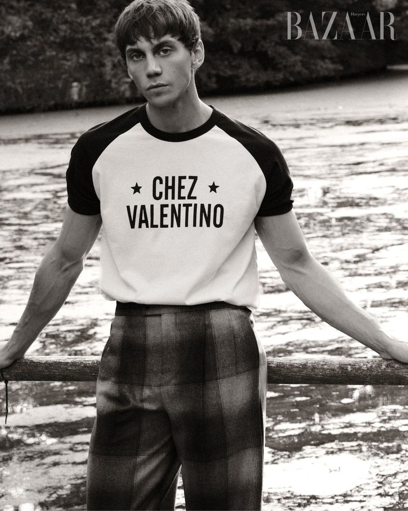 Black and white fashion shot of a young adult man in a t-shirt and plaid pants, leaning against a rail in a natural outdoor environment.