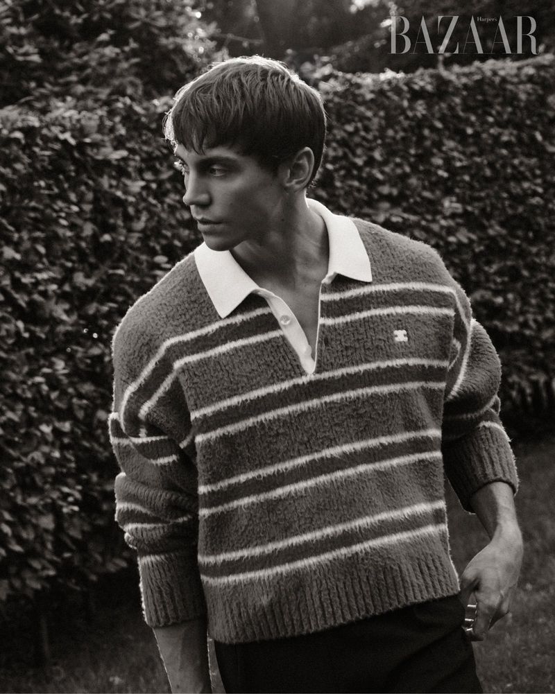 Black and white portrait of a young man in a striped sweater, standing outdoors with a hedge and 'BAZAAR' logo in the background.