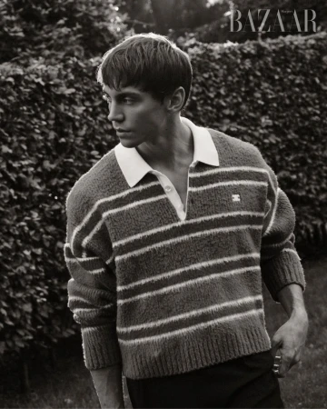 Black and white portrait of a young man in a striped sweater, standing outdoors with a hedge and 'BAZAAR' logo in the background.