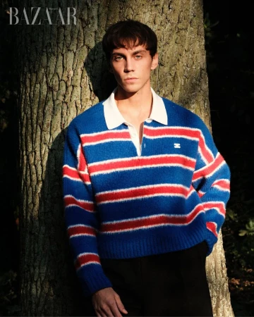 Matteo Miccini poses against a tree wearing a blue, red, and white striped collared knit sweater and black trousers.