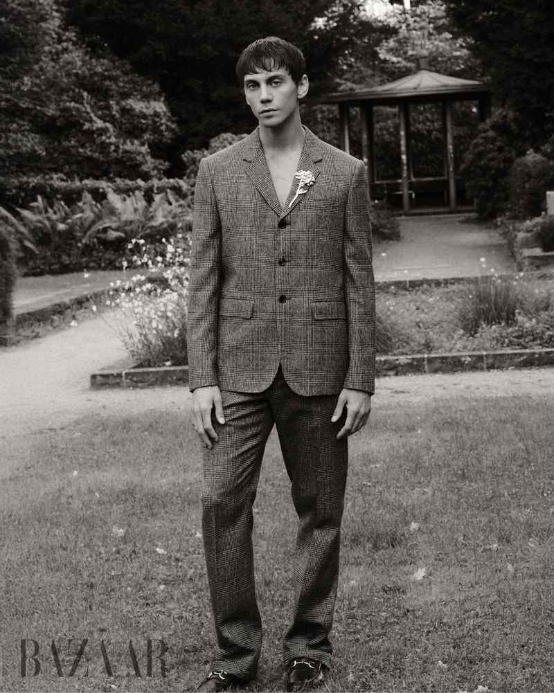 A man poses in a tweed suit and flower boutonniere in a park setting with a gazebo, captured in black and white for a vintage fashion editorial.