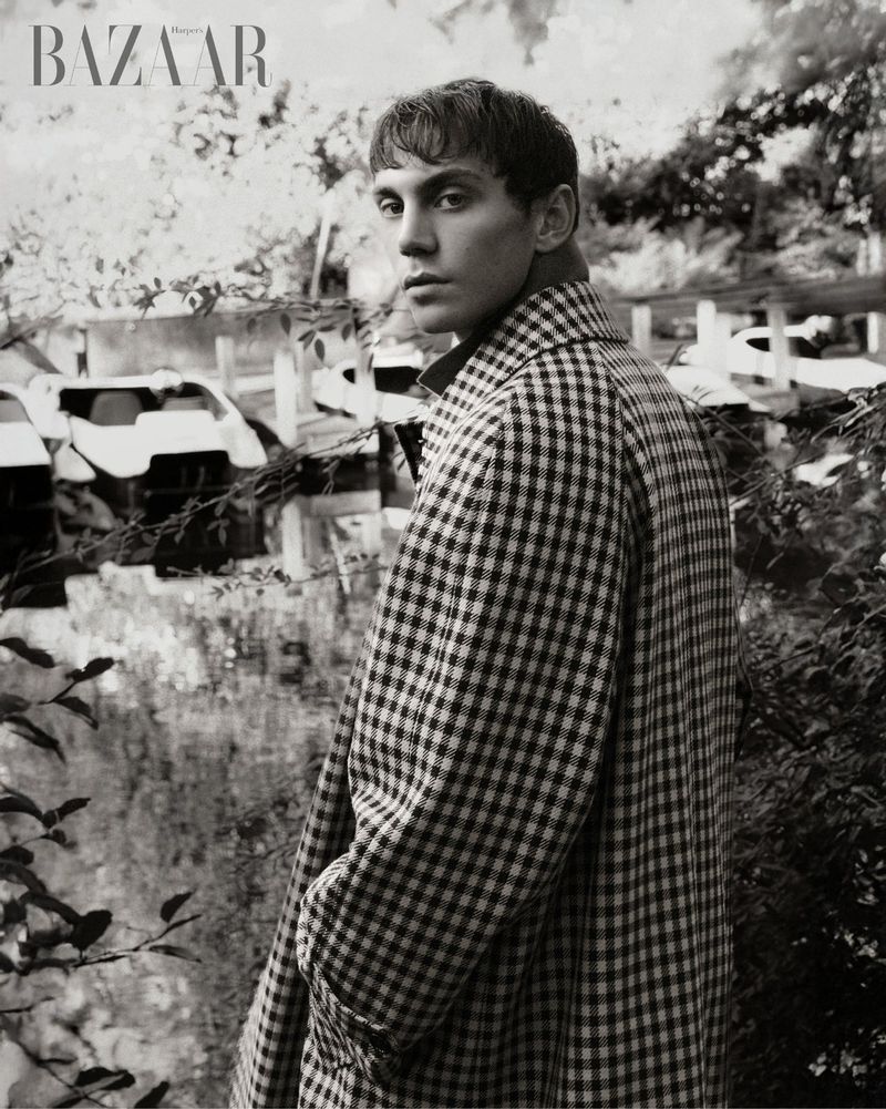 A young adult man gazes over his shoulder in a black and white portrait, wearing a checkered coat with blurred cars in the background.