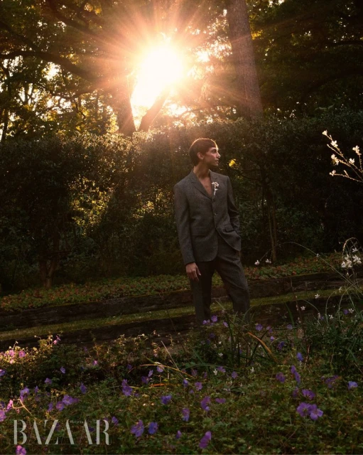 An adult male in a suit stands in a meadow surrounded by trees, plants, and purple wildflowers, bathed in sunlight for a warm and natural outdoor portrait.