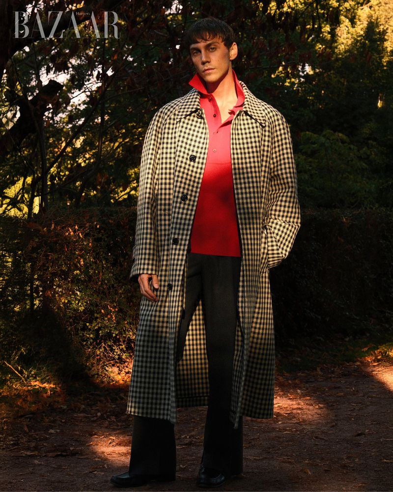 A male fashion model in a long, checkered coat and red polo shirt poses outdoors in a forest setting, styled for an editorial fashion shoot.