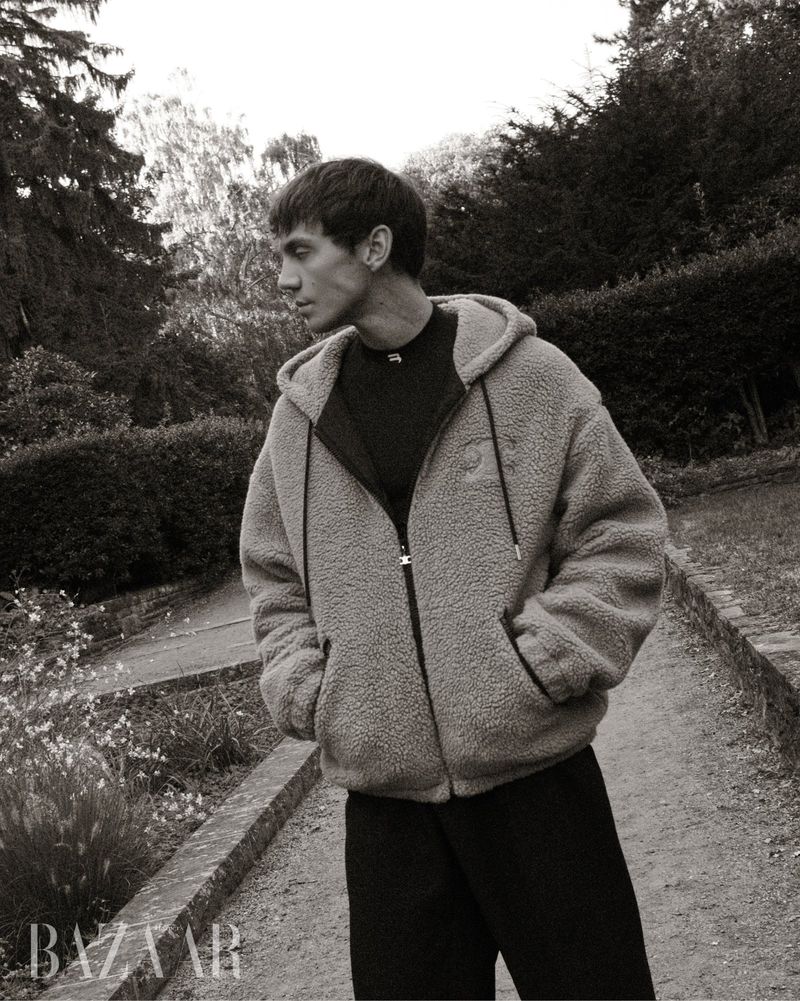 Matteo Miccini wearing a fleece zip-up hooded jacket and dark trousers standing on an outdoor garden path in monochrome.