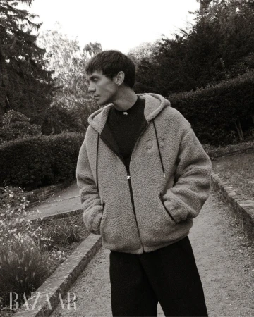 Matteo Miccini wearing a fleece zip-up hooded jacket and dark trousers standing on an outdoor garden path in monochrome.