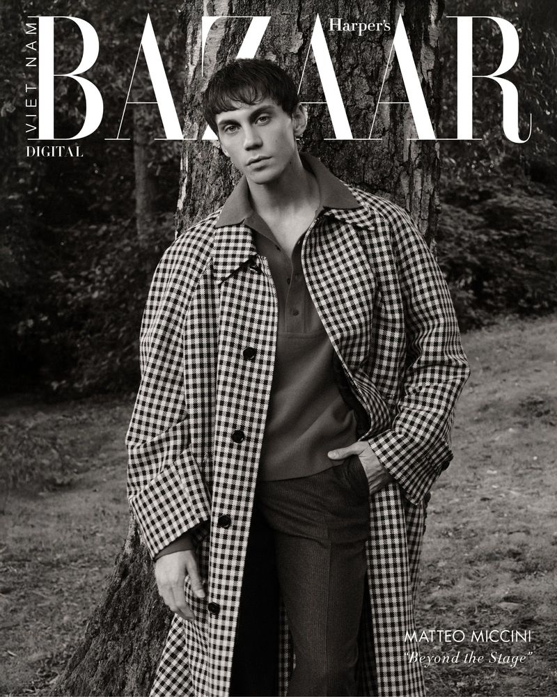 Matteo Miccini wearing a checkered overcoat and polo shirt while leaning against a tree in a woodland setting.