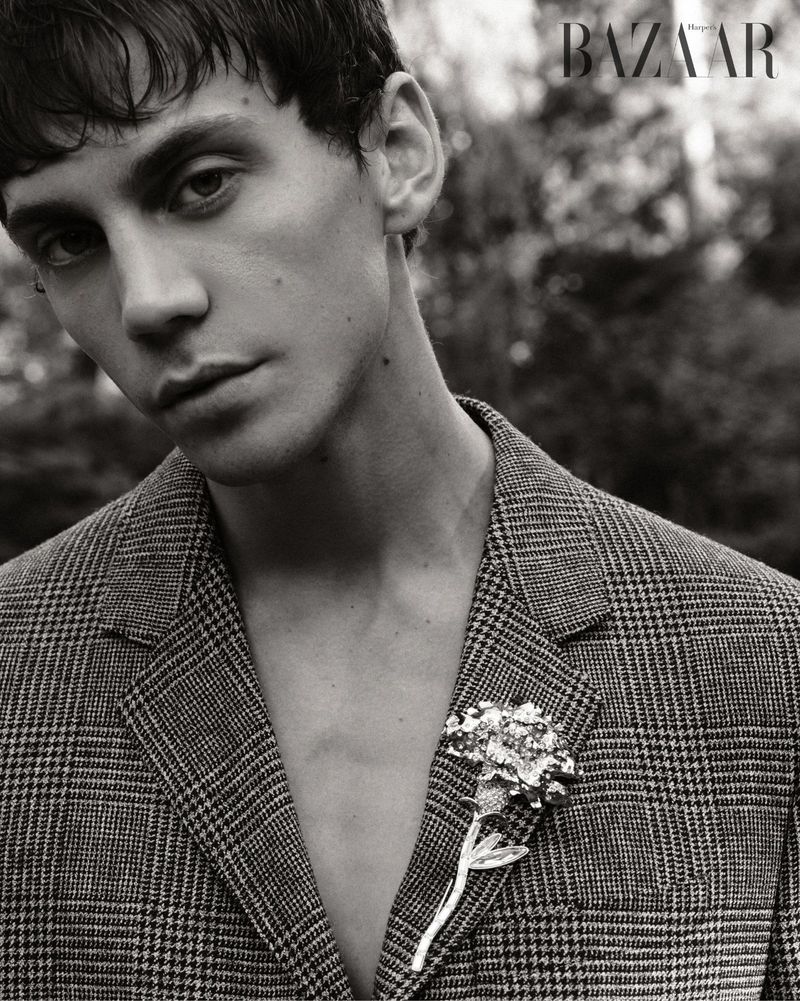 Monochrome portrait of a young adult man in a glen plaid jacket with a floral brooch, gazing at the viewer. Magazine logo visible.