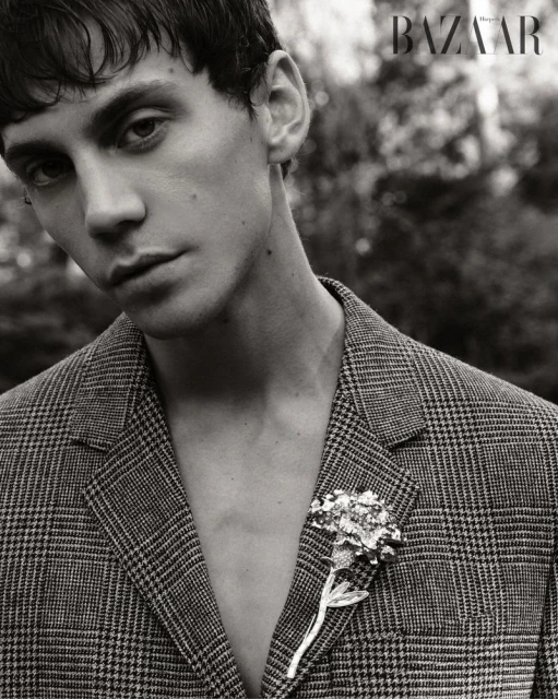 Monochrome portrait of a young adult man in a glen plaid jacket with a floral brooch, gazing at the viewer. Magazine logo visible.