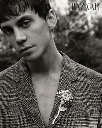Monochrome portrait of a young adult man in a glen plaid jacket with a floral brooch, gazing at the viewer. Magazine logo visible.
