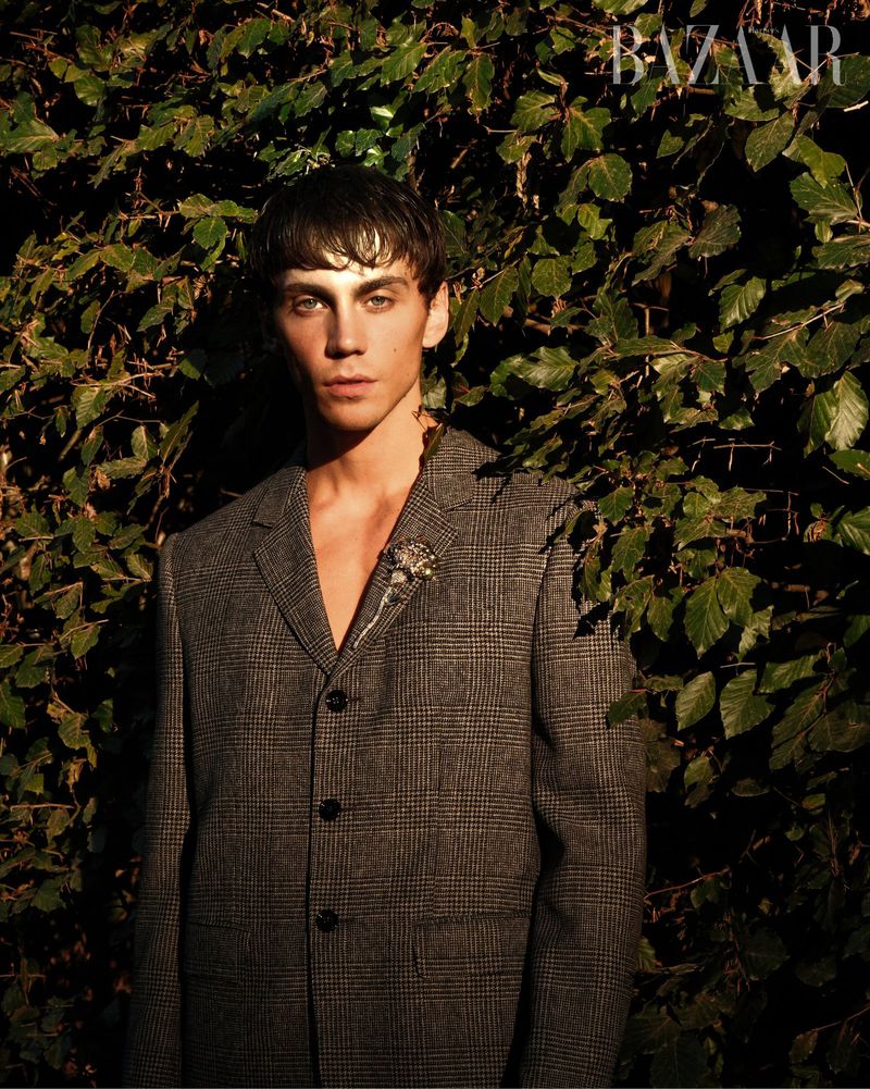 Portrait of a young adult male wearing a grey blazer, standing in front of a green leafy wall with soft, natural lighting.