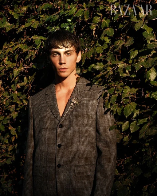 Portrait of a young adult male wearing a grey blazer, standing in front of a green leafy wall with soft, natural lighting.