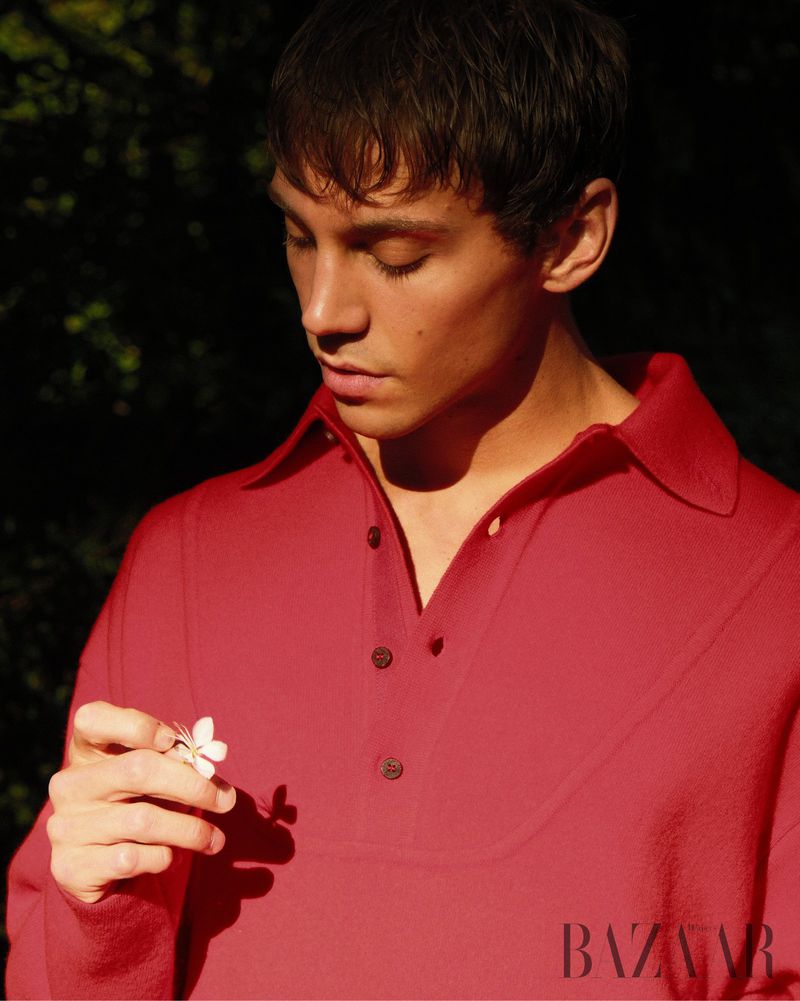 Matteo Miccini looking down while holding a small white flower, wearing a red collared knit polo shirt in direct sunlight.