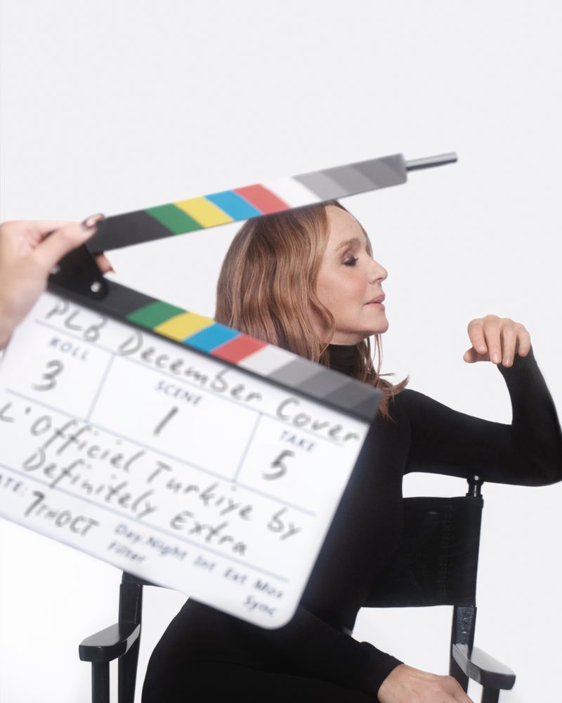 A woman with medium-length hair poses next to a clapperboard that displays 'December Cover' against a white background in a studio setting.