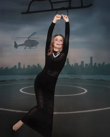 Woman in a black dress hangs from a black metal frame against a cityscape backdrop with a helicopter, posing playfully in a studio setting.