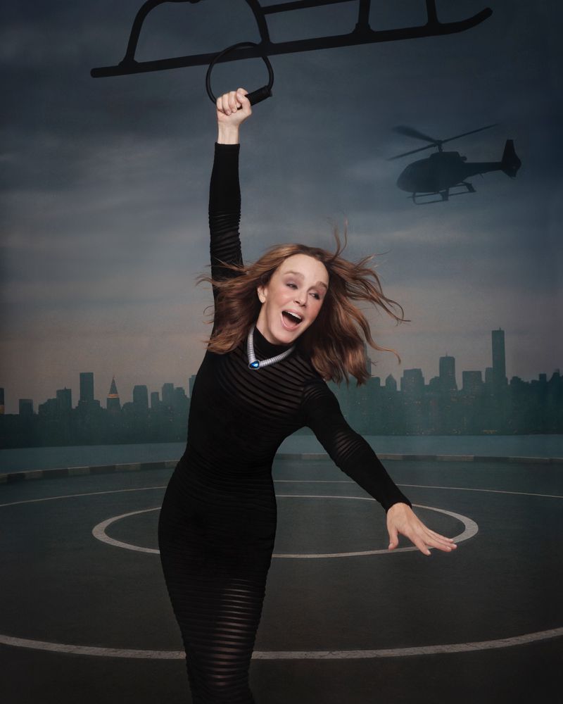 A woman in a black dress joyfully poses with a helicopter rig on a rooftop against a city skyline and cloudy sky background.