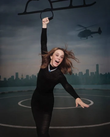 A woman in a black dress joyfully poses with a helicopter rig on a rooftop against a city skyline and cloudy sky background.
