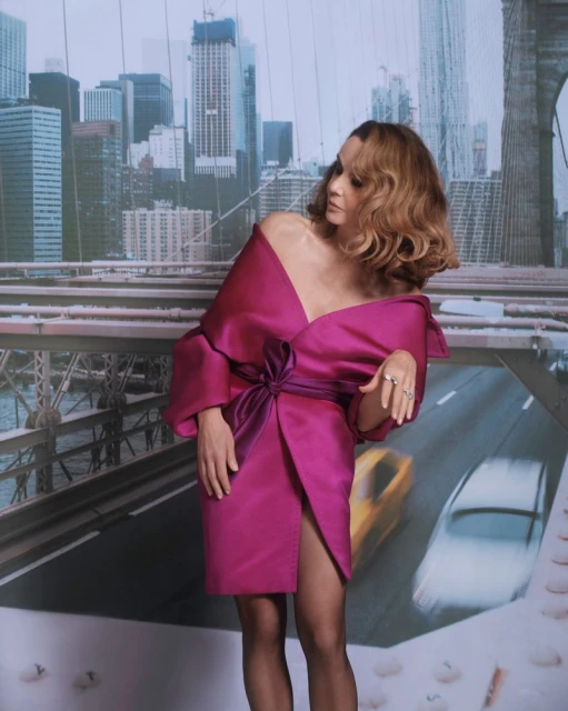 A woman in a magenta dress poses in front of a backdrop of the Brooklyn Bridge and NYC skyline, creating a fashion-forward studio portrait.