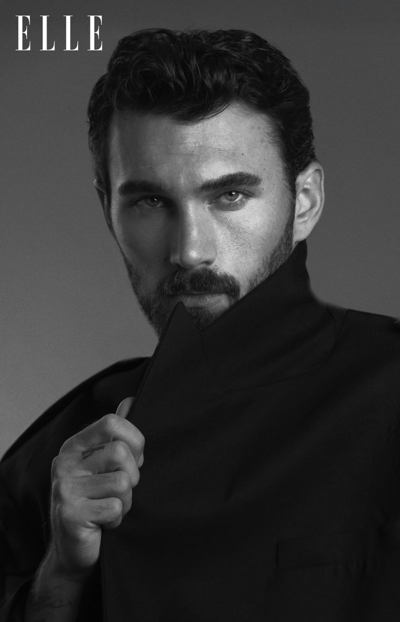 Black and white portrait of an adult male with dark hair and a beard, wearing a dark coat and standing in a studio setting.
