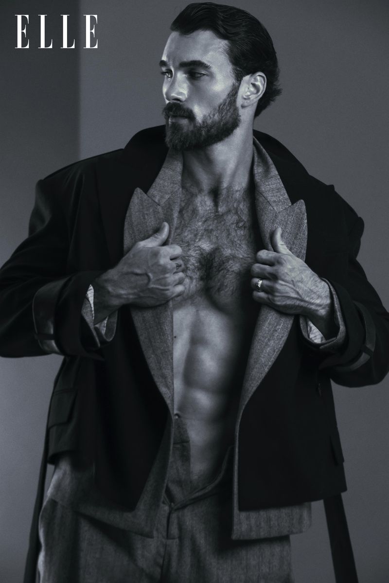 Monochrome studio portrait of a shirtless man with a beard, wearing a jacket and pants, looking off-camera near the 'ELLE' logo.
