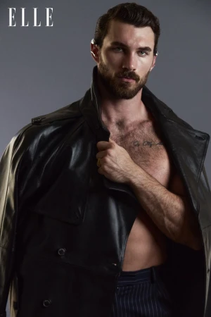 Male model with beard and black leather coat poses against gray backdrop; fashion editorial for Elle magazine, studio lighting.