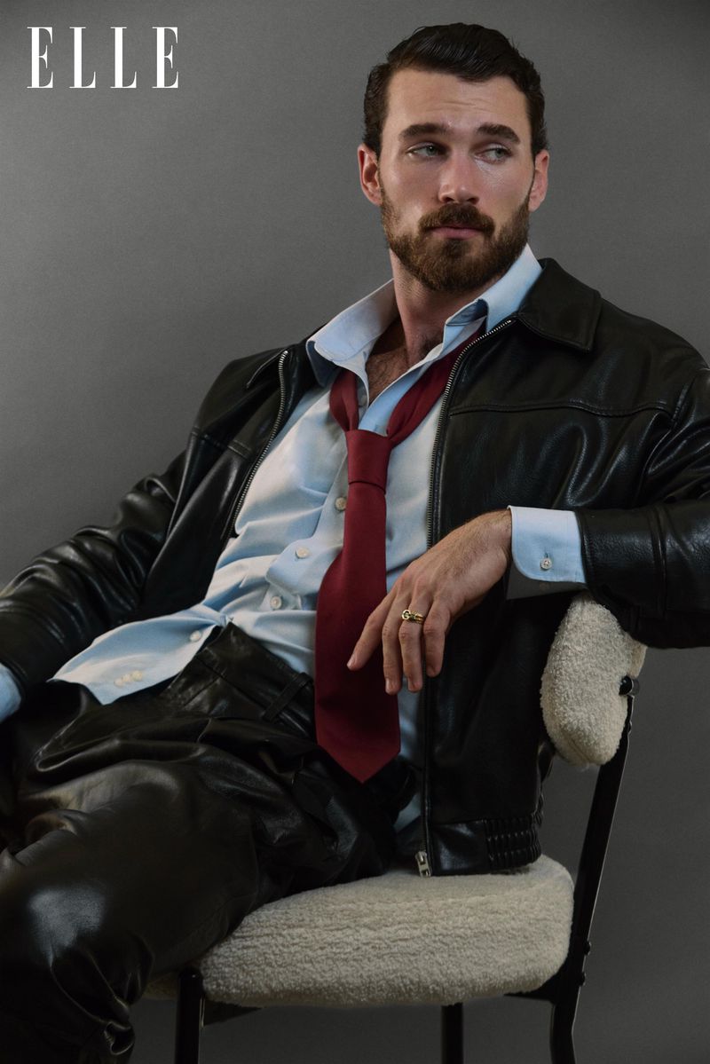 Portrait of a bearded man in a black leather jacket and a red tie, sitting on a chair against a grey backdrop, magazine cover.