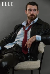 Model Michael Yerger seated on a white chair wearing a black leather jacket, light blue shirt, and red tie against a grey background.