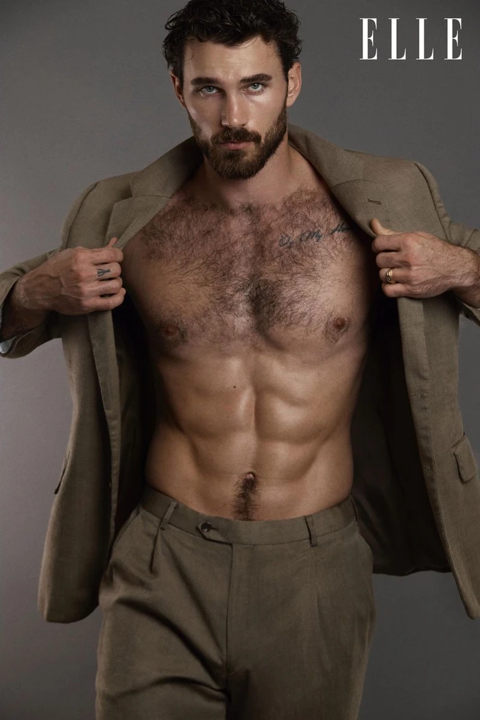 Model Michael Yerger posing shirtless in an open taupe suit jacket and tailored trousers against a grey backdrop.