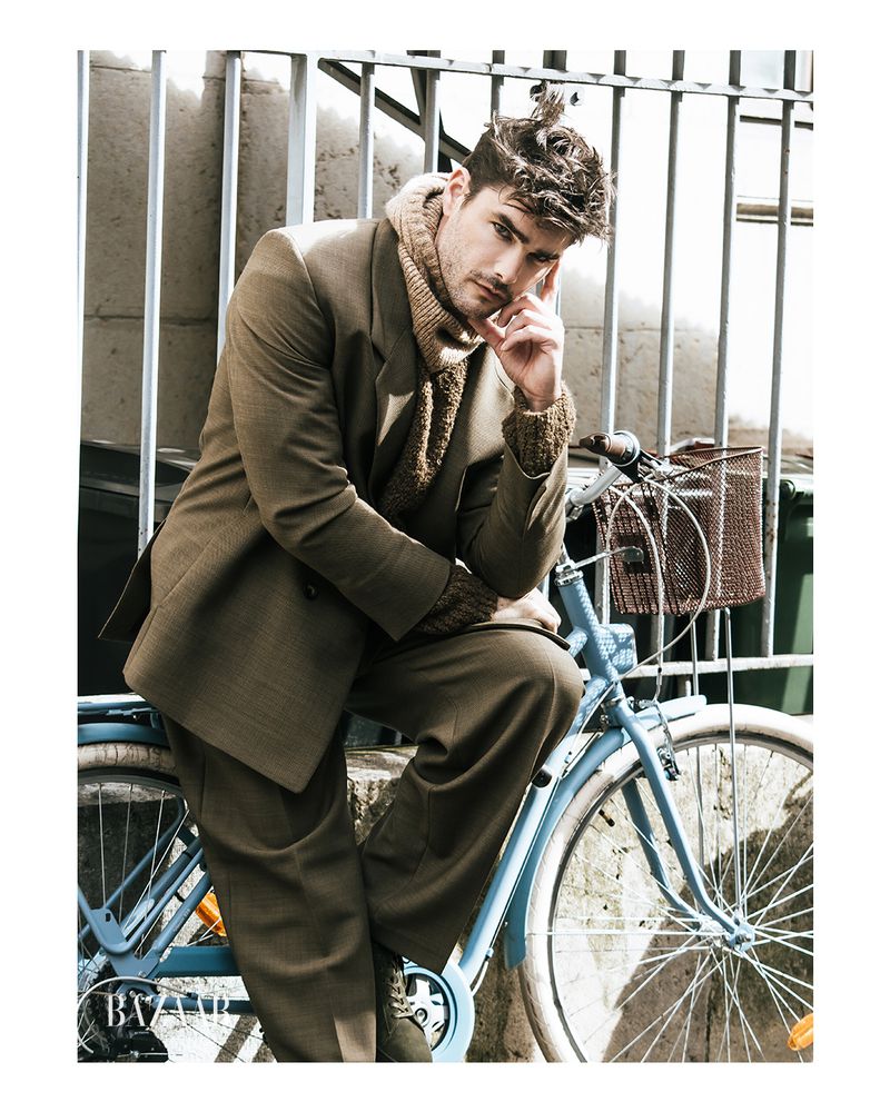 A man in a suit is sitting on a light blue bicycle in front of a metal fence outdoors, modelling in a fashion editorial shot.