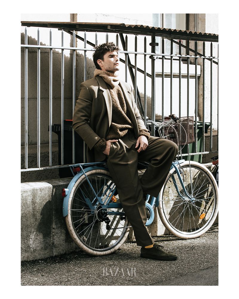 Adult male fashion model poses, wearing a suit and sweater, sitting on a light blue bicycle, against a metal railing outdoors.