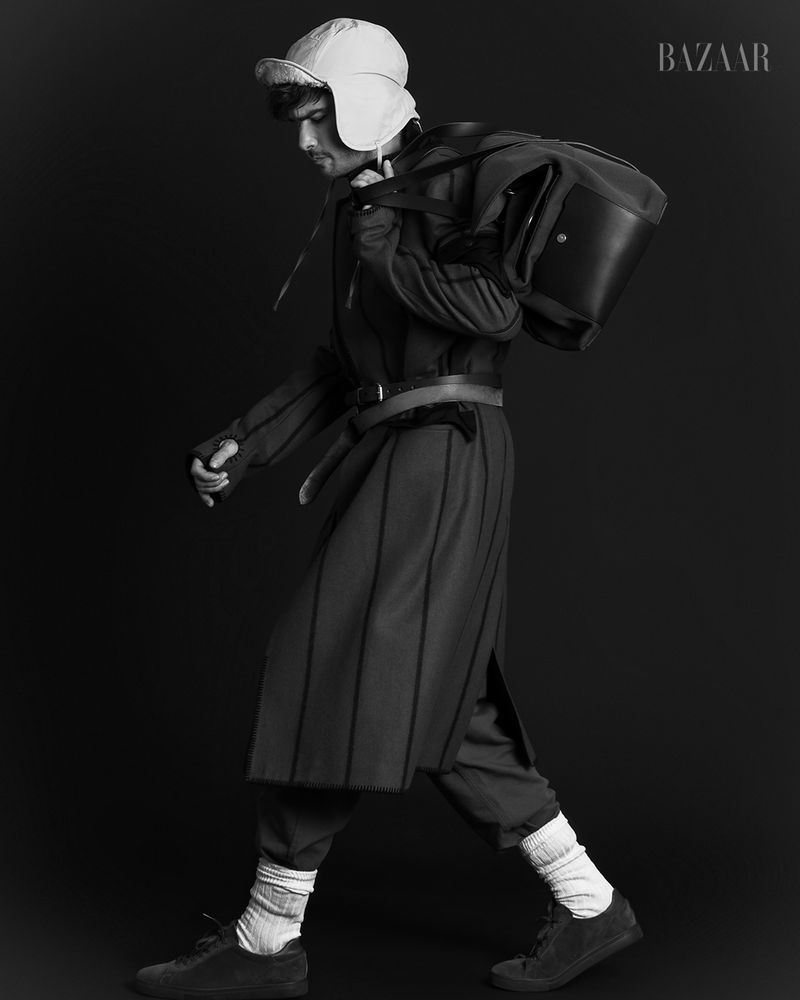 Fashion editorial shot in black and white features an adult male walking with a bag over his shoulder in a studio. He wears a hat.