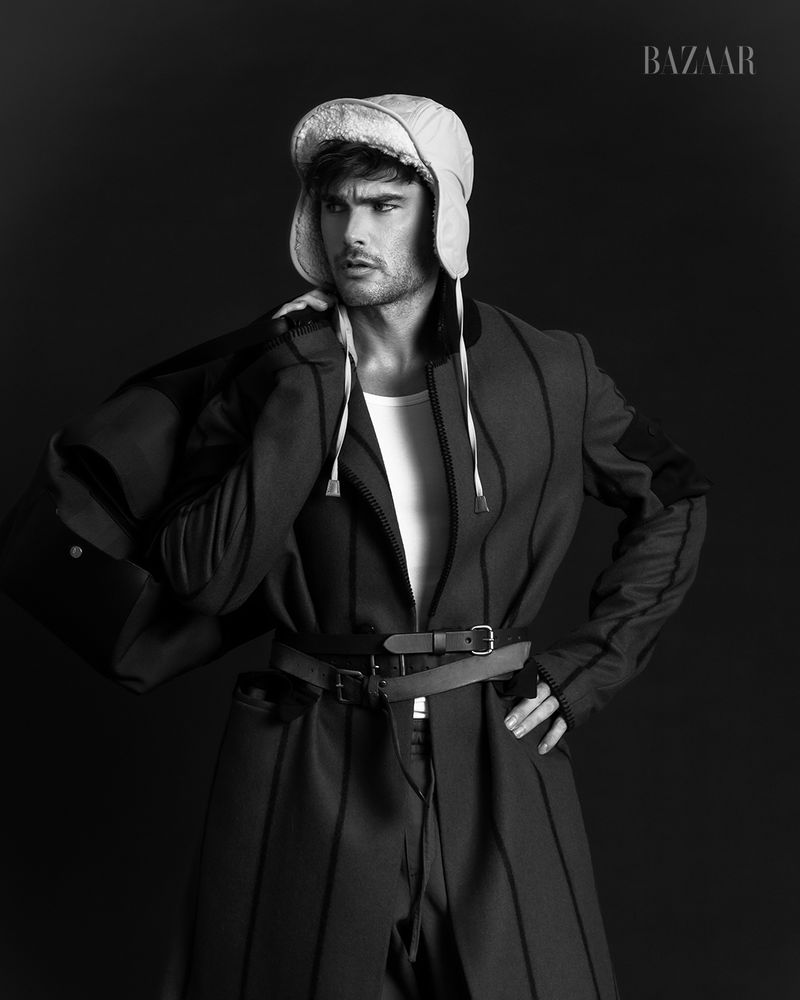 Black and white portrait of a male model in a heavy coat and hat, posing against a dark backdrop for a fashion editorial.