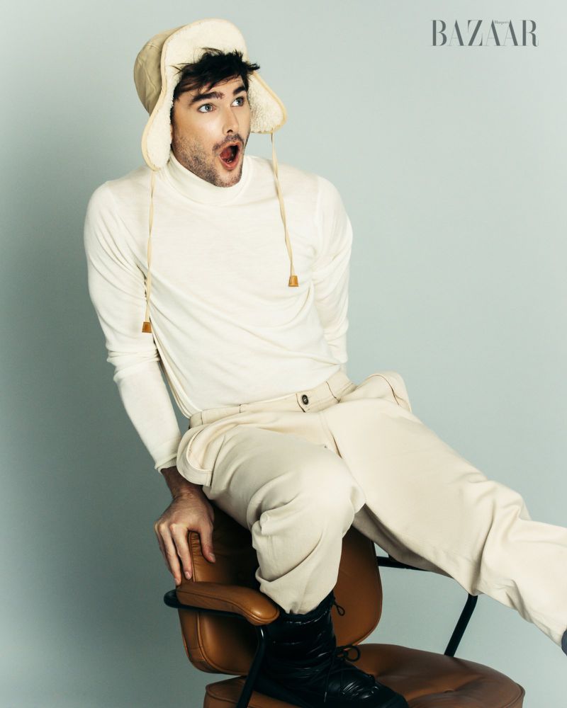 A man poses in a studio, wearing a cream-colored turtleneck, matching pants, and a fur-lined hat, seated on a brown chair against a light blue backdrop.