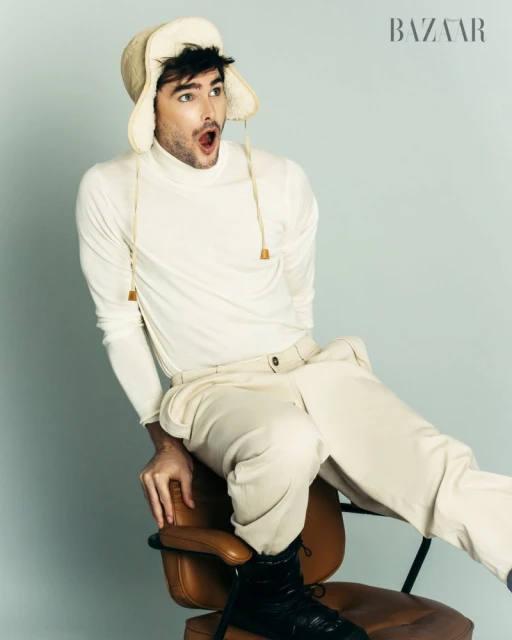 A man poses in a studio, wearing a cream-colored turtleneck, matching pants, and a fur-lined hat, seated on a brown chair against a light blue backdrop.