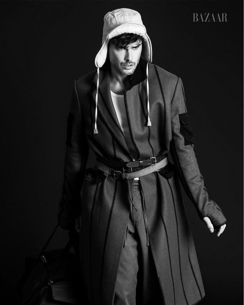 Black and white fashion editorial shot of a male model wearing a coat with vertical stripes, pants, and a trapper hat on black background.
