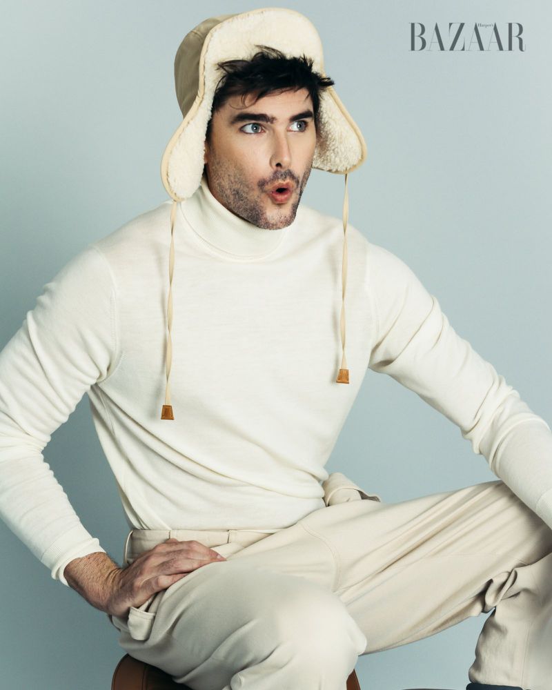 A fashionable adult male poses in a studio wearing a cream-colored turtleneck, pants, and fur-lined trapper hat against a light gray background.