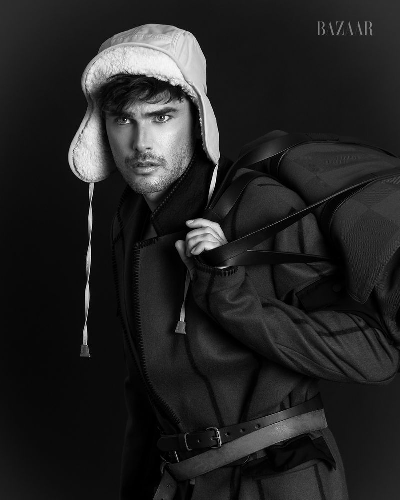 Black and white portrait of a dark-haired adult male wearing a trapper hat and coat, carrying a large duffel bag over his shoulder.