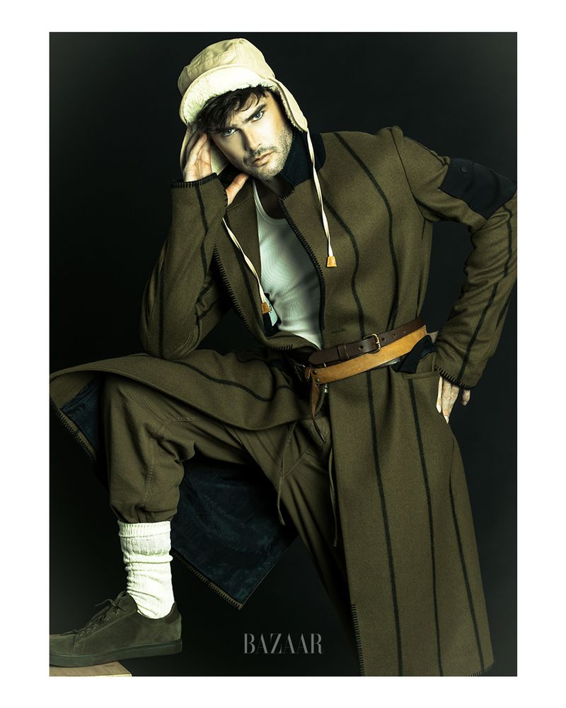 A fashionable adult male in a tan hat and striped brown coat poses in a photo studio against a black background for a Bazaar fashion editorial.