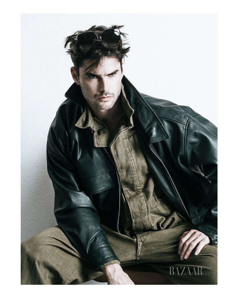 A tanned adult male model poses indoors wearing a black leather jacket, tan shirt and pants, and sunglasses, in front of a white background.