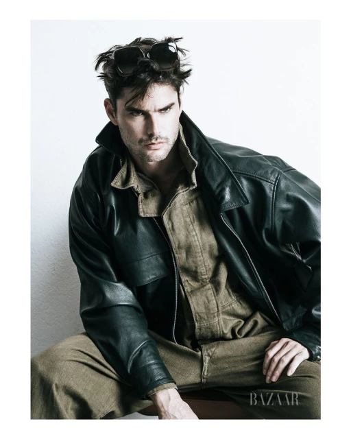 A tanned adult male model poses indoors wearing a black leather jacket, tan shirt and pants, and sunglasses, in front of a white background.