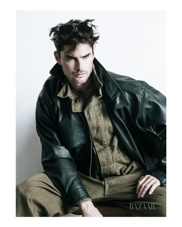 Model Charlie Matthews in an oversized leather jacket and olive jumpsuit with sunglasses on his head against a white background.