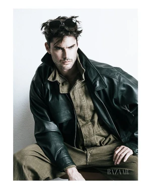Model Charlie Matthews in an oversized leather jacket and olive jumpsuit with sunglasses on his head against a white background.
