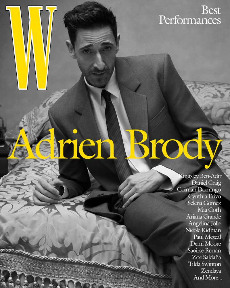 Adrien Brody poses in a grey suit for a W Magazine cover, overlaid with the magazine's title and other text, in a gray-toned photo.