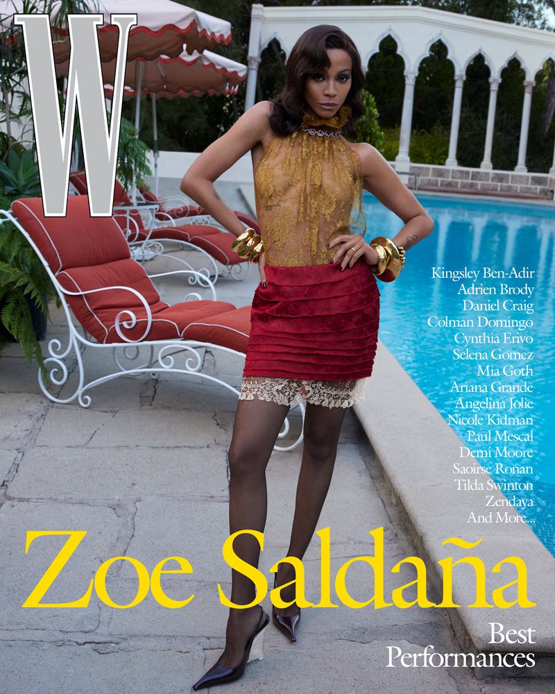 Zoe Saldana poses in a gold top and red skirt for a magazine cover in an outdoor setting with lounge chairs and a pool.