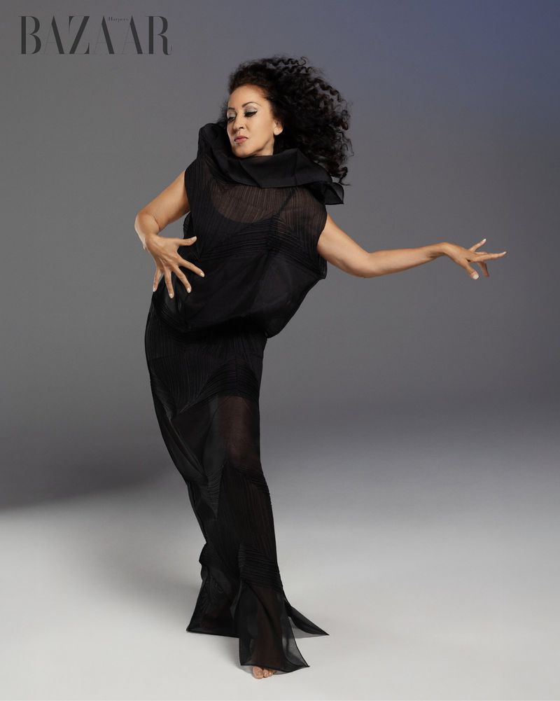 Woman poses in a pleated black dress with a high collar against a gray background, fashion editorial, studio lighting, and magazine logo.