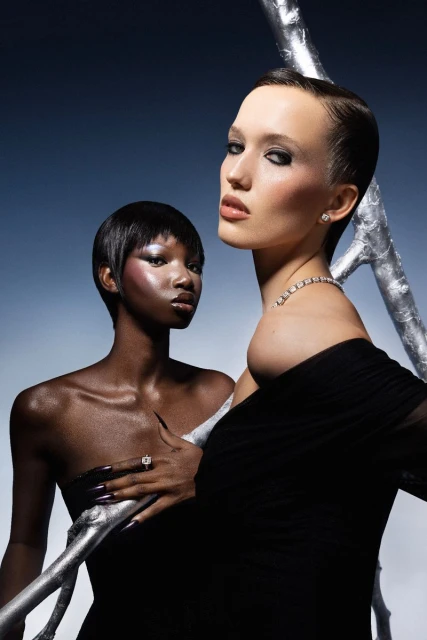 Two women with short hair pose in front of a silver metallic structure against a dark blue backdrop, wearing black dresses and jewelry.