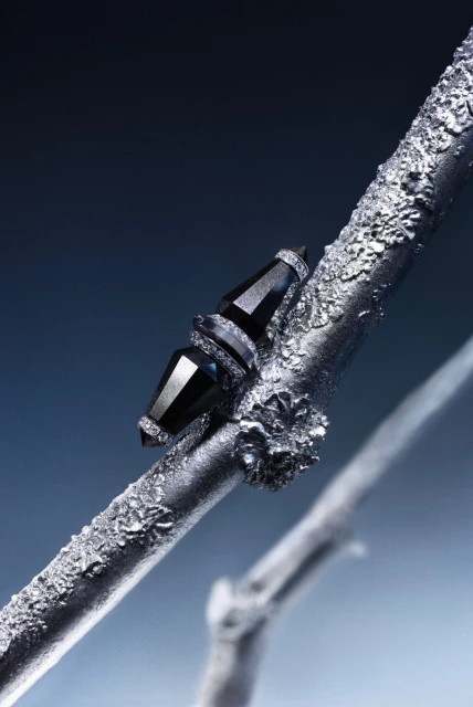 Close-up of black gemstone jewelry set on a textured silver branch against a gradient dark blue background in vertical format.