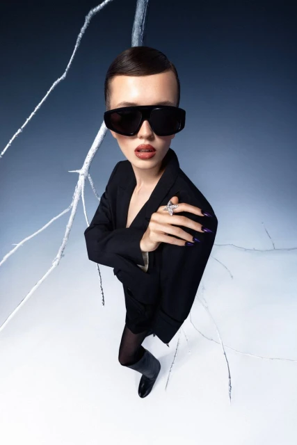 Stylish woman in black blazer and sunglasses poses with nail art, ring, and branches in a high-angle fashion studio portrait.